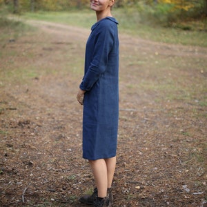 May include: A woman wearing a long-sleeved, knee-length navy blue dress with a collar. The dress is made of a solid fabric. She is wearing brown boots and standing on a path in a forest.