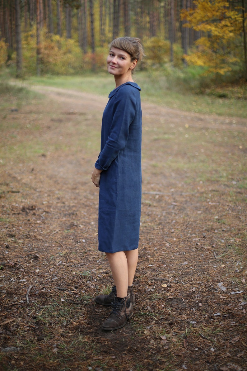 May include: A woman wearing a long-sleeved, knee-length navy blue dress with a collar. The dress is made of a solid fabric. She is wearing brown boots and standing on a path in a forest.