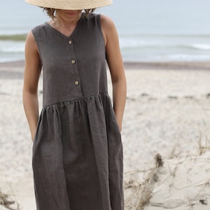 May include: A woman wearing a brown linen dress with a button-up front and pockets. She is wearing a wide-brimmed straw hat and is standing on a sandy beach.