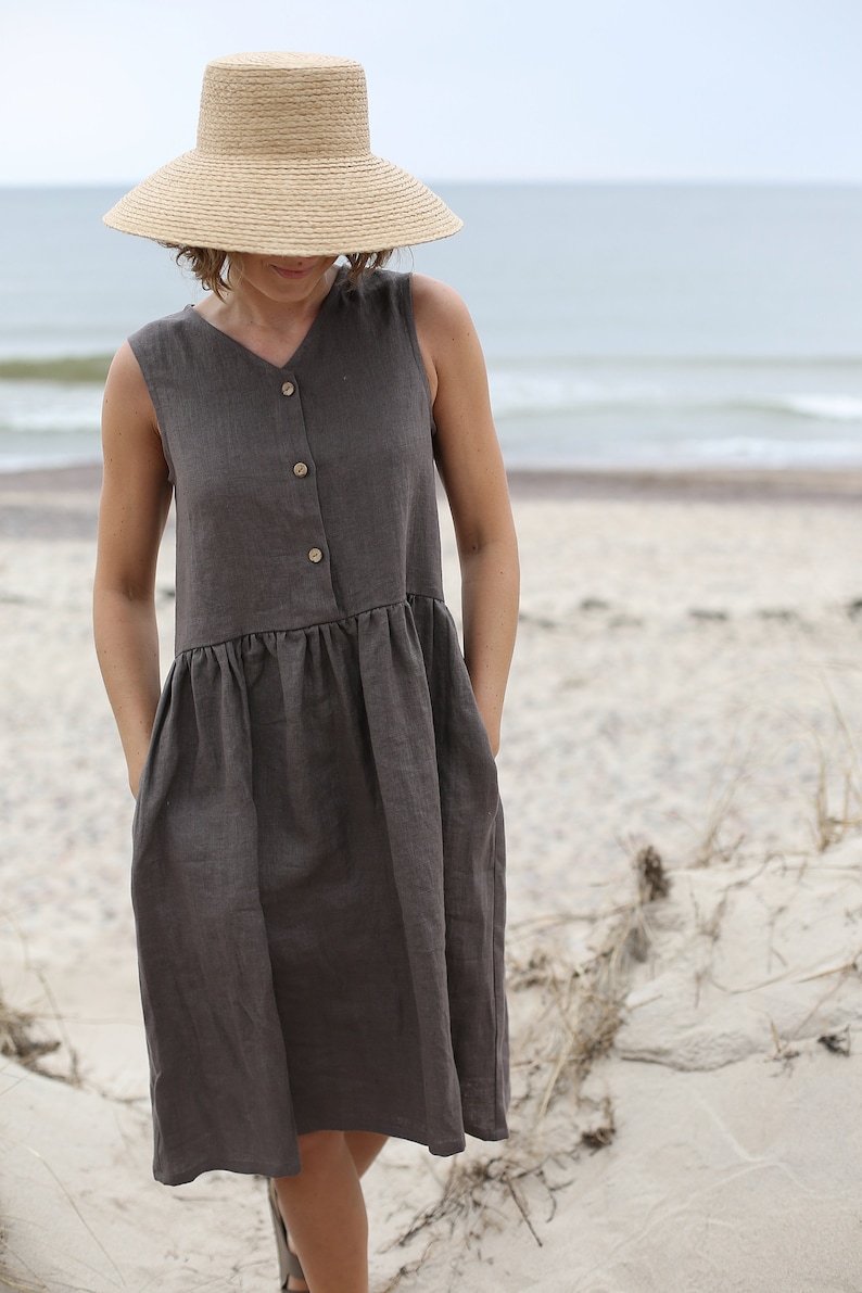 May include: A woman wearing a brown linen dress with a button-up front and pockets. She is wearing a wide-brimmed straw hat and is standing on a sandy beach.