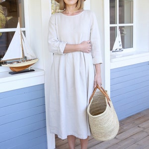 Pu&ograve; includere: Una donna indossa un abito di lino beige chiaro con un cappello di paglia e sandali neri. Tiene una borsa di vimini con manici in pelle.