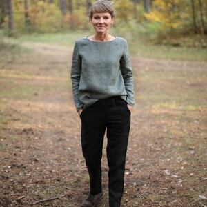 May include: A woman wearing a sage green linen top and black pants stands in a forest setting. She is looking at the camera with a neutral expression. The background is blurred and shows trees with fall foliage.