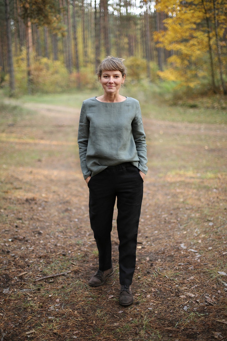 May include: A woman wearing a sage green linen top and black pants stands in a forest setting. She is looking at the camera with a neutral expression. The background is blurred and shows trees with fall foliage.