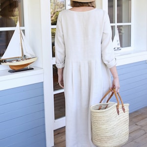 Pu&ograve; includere: Una donna indossa un abito di lino beige con un cappello di paglia e un nastro nero. Tiene una borsa di vimini con manici in pelle.
