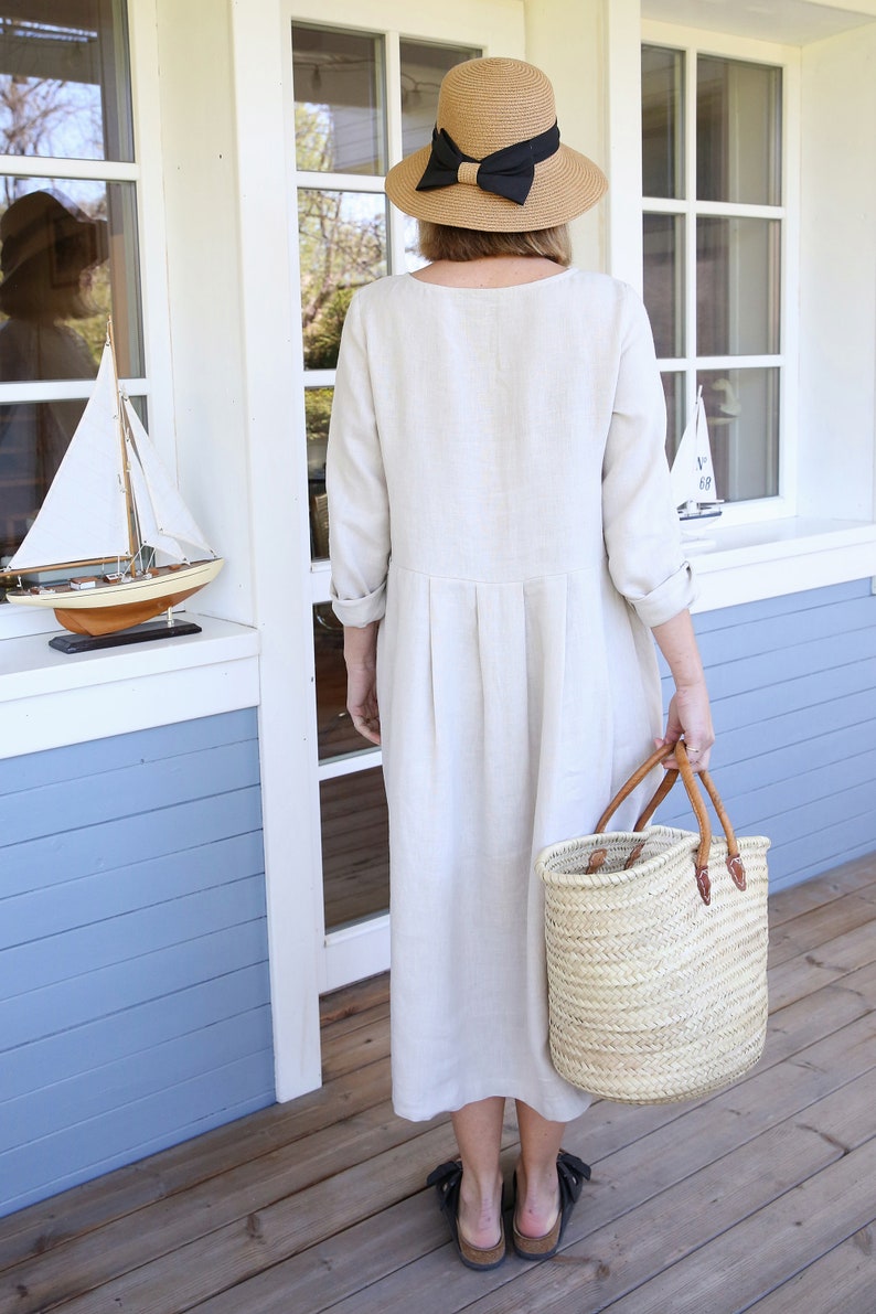 Pu&ograve; includere: Una donna indossa un abito di lino beige con un cappello di paglia e un nastro nero. Tiene una borsa di vimini con manici in pelle.