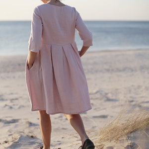 May include: A woman wearing a light pink linen dress with a pleated skirt and 3/4 sleeves stands on a sandy beach with tall grass in the background.