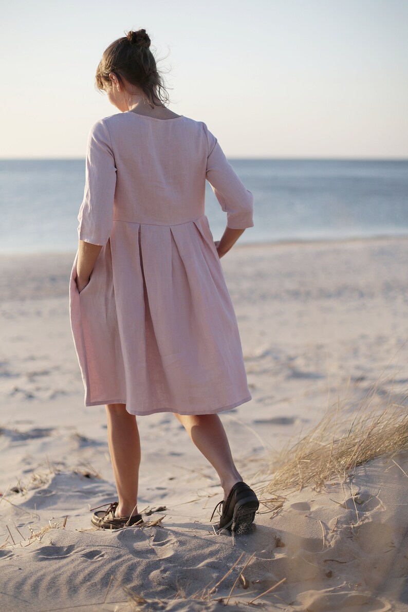 May include: A woman wearing a light pink linen dress with a pleated skirt and 3/4 sleeves stands on a sandy beach with tall grass in the background.