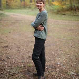 May include: A woman wearing a green long-sleeved shirt and black pants stands in a forest setting. She is smiling and looking at the camera.