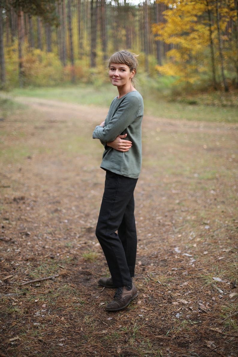 May include: A woman wearing a green long-sleeved shirt and black pants stands in a forest setting. She is smiling and looking at the camera.