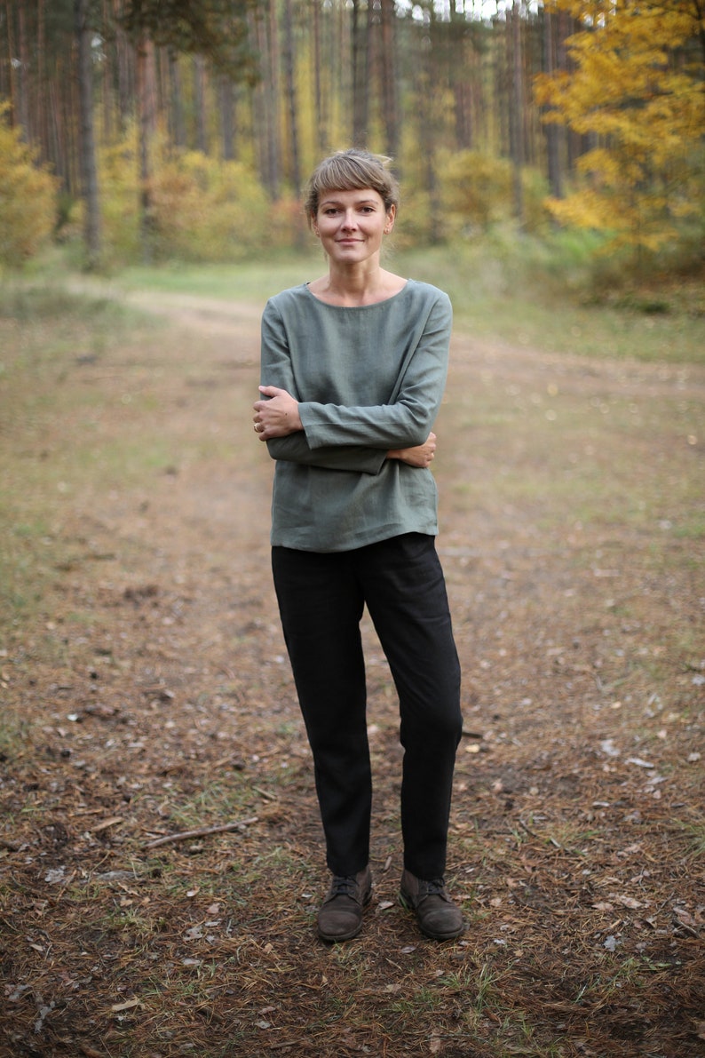 May include: A woman wearing a green long-sleeved shirt and black pants stands in a forest setting. The trees are in the background and the ground is covered in pine needles.