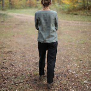 May include: A person wearing a green linen shirt and black pants walks on a path in a forest. The path is covered in pine needles and leaves.
