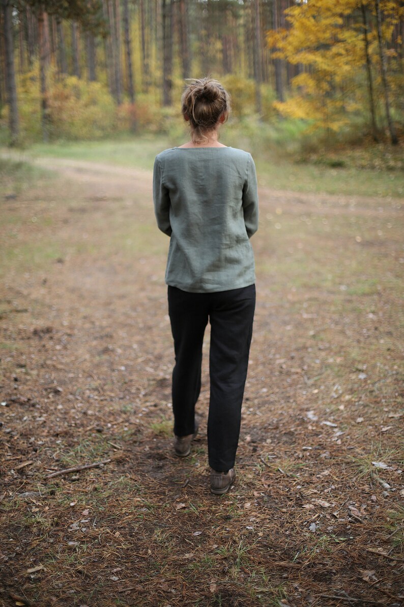 May include: A person wearing a green linen shirt and black pants walks on a path in a forest. The path is covered in pine needles and leaves.