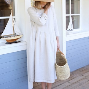 Pu&ograve; includere: Una donna indossa un abito di lino beige chiaro con un cappello di paglia e sandali neri. Tiene un cesto intrecciato.