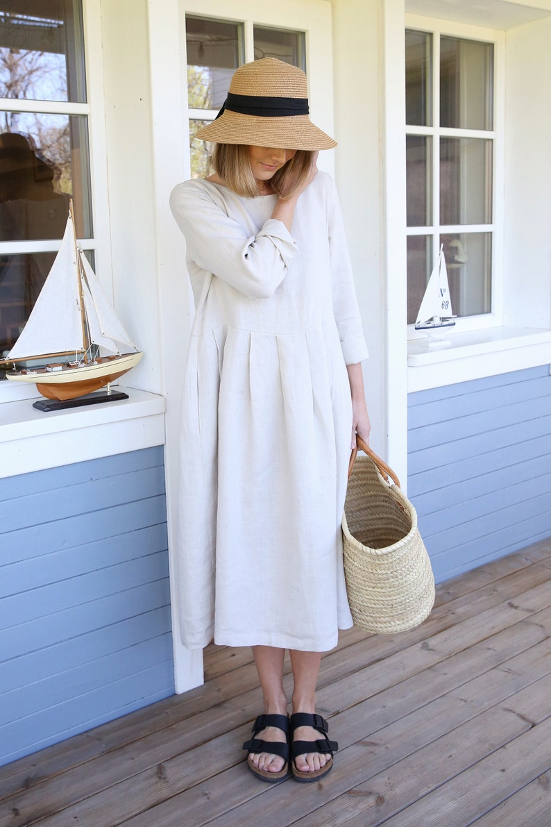Pu&ograve; includere: Una donna indossa un abito di lino beige chiaro con un cappello di paglia e sandali neri. Tiene un cesto intrecciato.