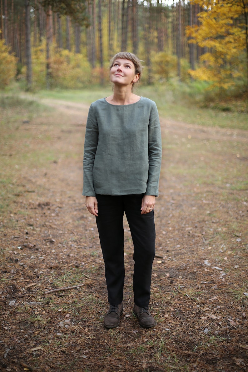 May include: A woman wearing a green linen shirt and black pants stands in a forest setting. She is looking up at the sky.