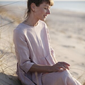 May include: A woman wearing a light pink linen dress sits in a grassy area with a sandy beach in the background.