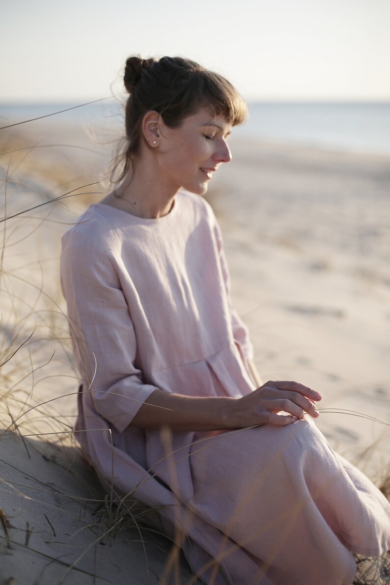 May include: A woman wearing a light pink linen dress sits in a grassy area with a sandy beach in the background.