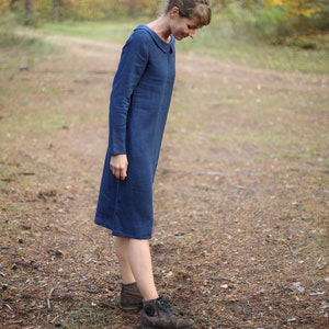 May include: A long-sleeved, knee-length blue dress with a rounded collar. The dress is made of a soft fabric and is worn with brown boots. The background features a forest with a dirt path.