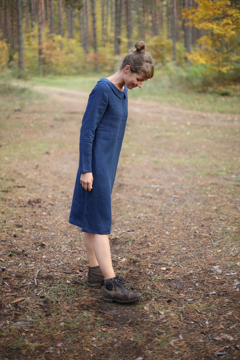 May include: A long-sleeved, knee-length blue dress with a rounded collar. The dress is made of a soft fabric and is worn with brown boots. The background features a forest with a dirt path.