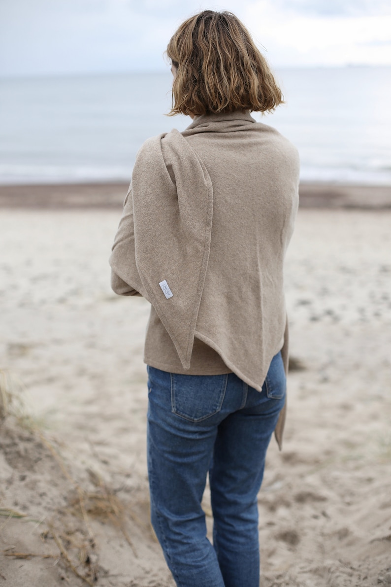 Może przedstawiać: Kobieta w beżowym szaliku kaszmirowym z białą metką z boku. Stoi na piaszczystej plaży z oceanem w tle.
