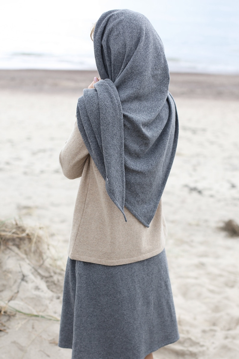 Może przedstawiać: Kobieta w szarym dzianym szaliku i beżowym swetrze z szarą sp&oacute;dnicą. Kobieta stoi na piaszczystej plaży.