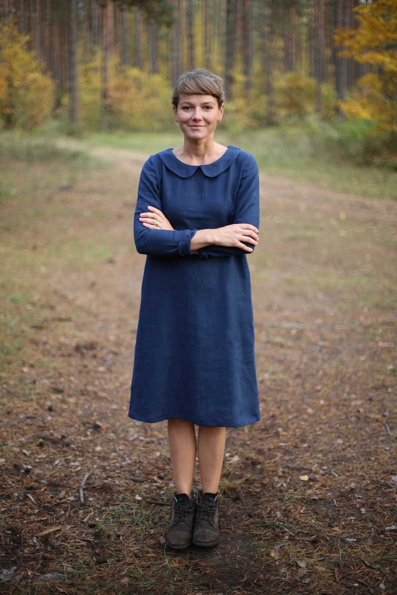May include: A woman wearing a long-sleeved, navy blue dress with a rounded collar. The dress falls to the mid-calf and is paired with dark brown ankle boots. The background is a blurred forest scene.