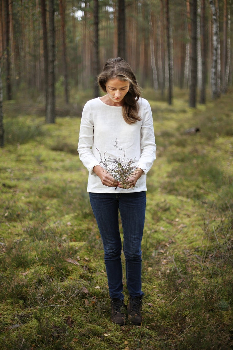 May include: A woman in a white long-sleeved shirt stands in a forest, holding a small bouquet of dried branches.