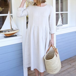 Pu&ograve; includere: Una donna indossa un abito di lino beige con un cappello di paglia e sandali neri. Tiene una borsa di vimini.