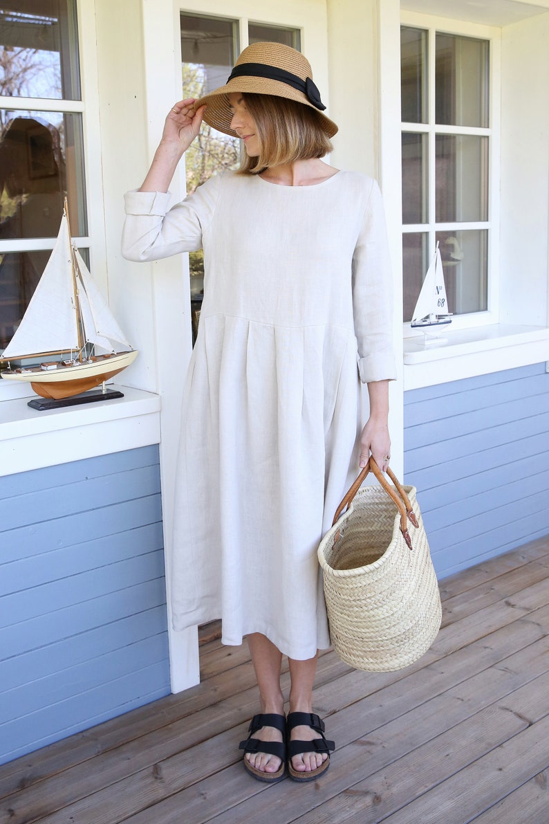 Pu&ograve; includere: Una donna indossa un abito di lino beige con un cappello di paglia e sandali neri. Tiene una borsa di vimini.