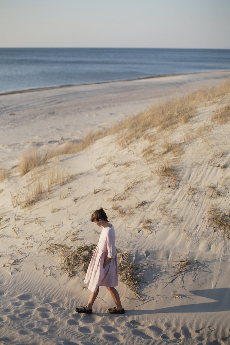 May include: A woman in a pink dress walks on a sandy beach with tall grass in the background.