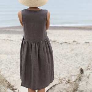 May include: A woman wearing a brown linen dress and a wide-brimmed straw hat stands on a sandy beach, facing the ocean.