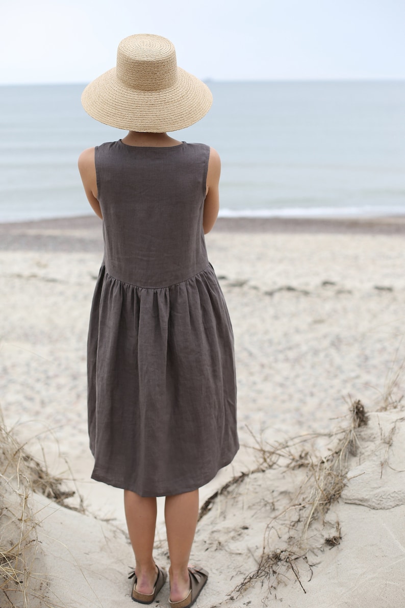May include: A woman wearing a brown linen dress and a wide-brimmed straw hat stands on a sandy beach, facing the ocean.