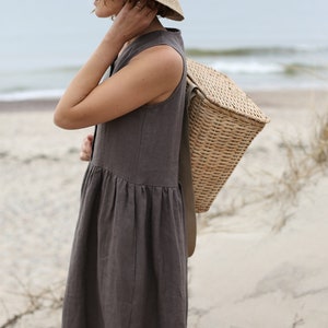 May include: A woman wearing a brown linen dress and a wide-brimmed straw hat carries a woven wicker basket on her shoulder.