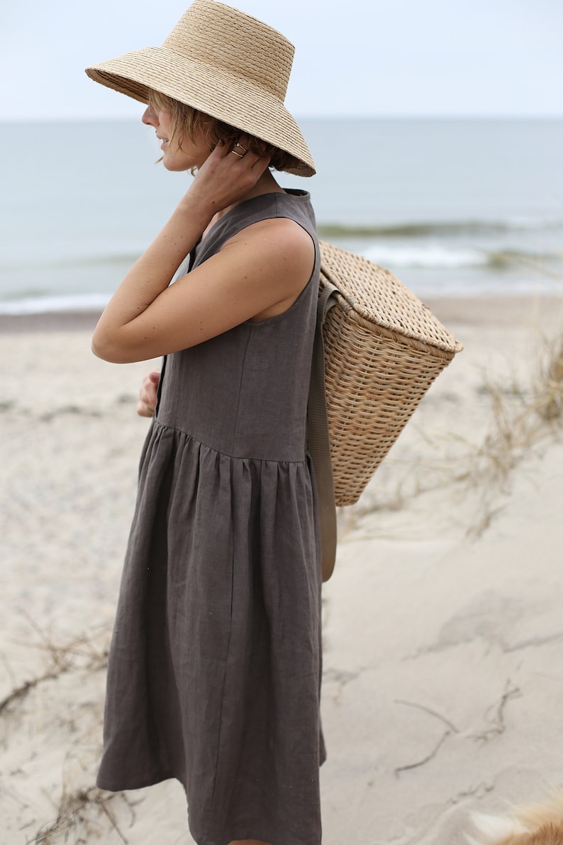May include: A woman wearing a brown linen dress and a wide-brimmed straw hat carries a woven wicker basket on her shoulder.