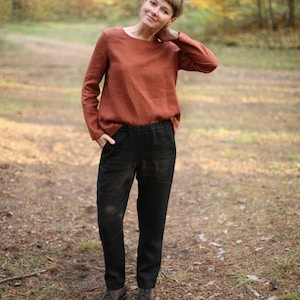 May include: A woman wearing a rust-colored linen top and black linen pants stands in a forest setting. She is looking at the camera and has her hand on her neck.
