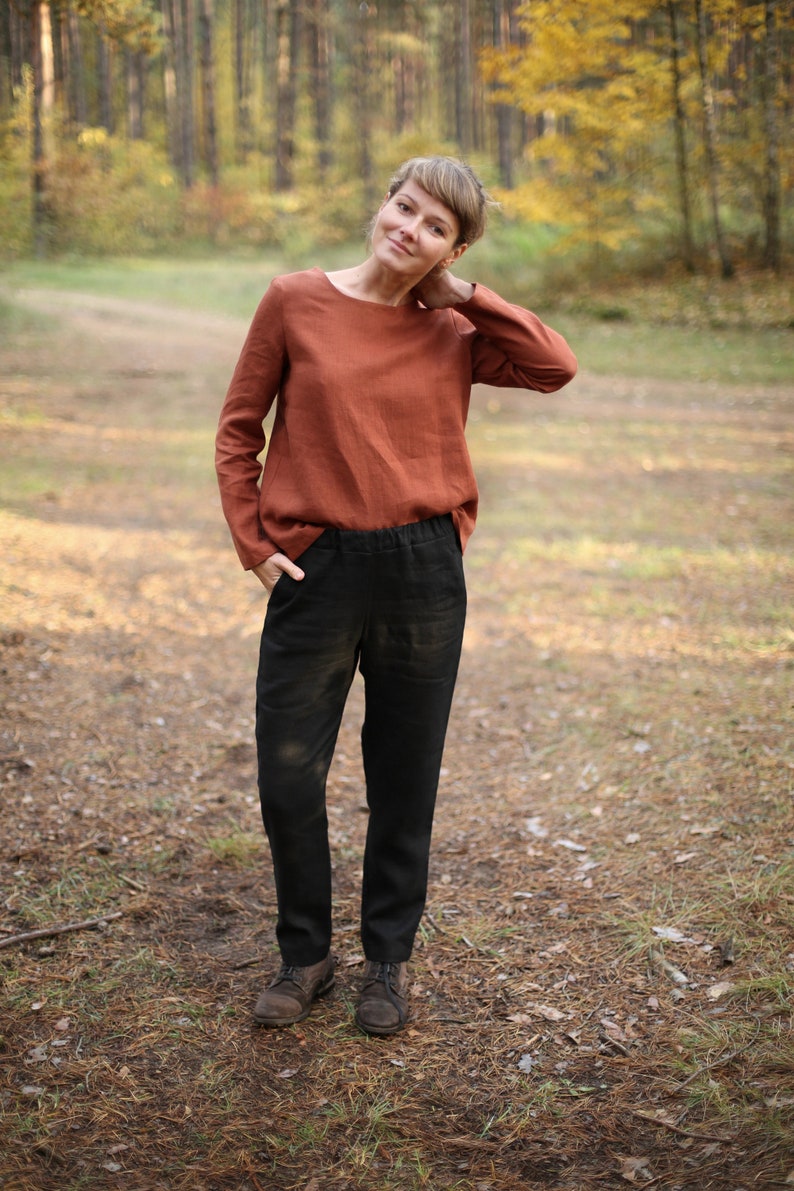 May include: A woman wearing a rust-colored linen top and black linen pants stands in a forest setting. She is looking at the camera and has her hand on her neck.
