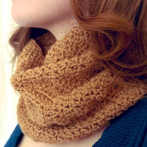 Könnte beinhalten: Nahaufnahme eines gehäkelten Infinity-Schals in einem warmen, goldbraunen Farbton. Der Schal ist um den Hals einer Person gewickelt, wobei die strukturierten Maschen deutlich sichtbar sind. Der Hintergrund umfasst ein blaues Oberteil und rotes Haar.