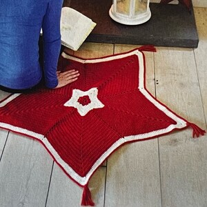 May include: A red crocheted star-shaped rug with a white border and a white crocheted star in the centre. The rug is on a wooden floor in front of a fireplace.