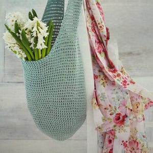 May include: A pale blue crocheted bag filled with white flowers. A floral scarf with pink and red roses hangs nearby. The bag is suspended from a white wooden structure, creating a soft, spring-like aesthetic.