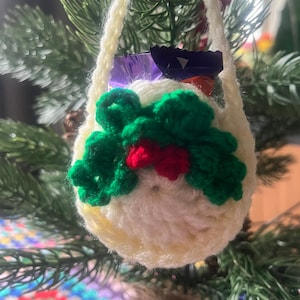 May include: A white crocheted bag with a green holly design and a red berry. The bag is filled with small wrapped candies and is hanging on a green Christmas tree.