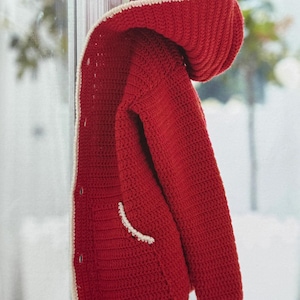 May include: A red crochet jacket with a hood and cream-coloured trim. The jacket is buttoned and hanging on a white hook. The crochet stitch is visible, and the hood is folded over. The jacket has a pocket.