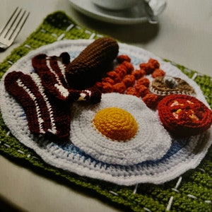 Puede incluir: Un plato de desayuno de ganchillo con un huevo frito, tocino, salchicha, frijoles horneados, champiñones y un tomate. El plato está sobre una alfombra de ganchillo verde.