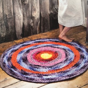 Può includere: Un tappeto rotondo all'uncinetto colorato con cerchi concentrici di filo viola, arancione, rosa e giallo. Il tappeto è su un pavimento di legno.