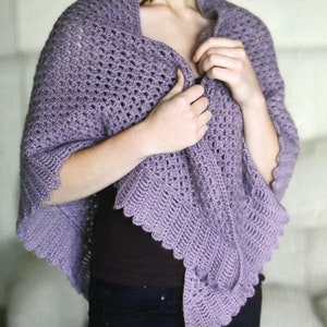May include: A lavender crochet shawl with a scalloped edge. The shawl is draped over a person's shoulders and is being held in place by their hands.