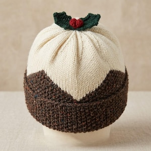 May include: A knitted hat designed to resemble a Christmas pudding. The hat features a cream-colored top with a brown base, and is topped with a green holly leaf and red berry embellishment. The hat is a festive accessory.