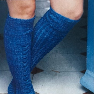 May include: A pair of blue knitted knee-high socks with a cable knit pattern. The socks are worn on a person's legs and the feet are pointed.