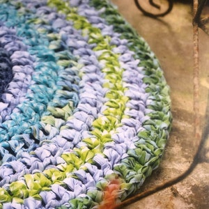 May include: A close-up of a round, crocheted rug with blue, green, and white stripes. The rug is made from recycled fabric and has a unique, textured look.