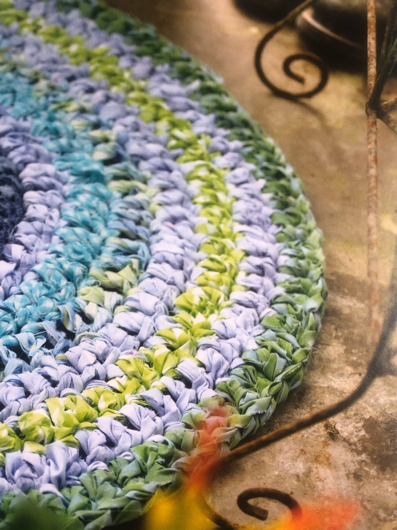 May include: A close-up of a round, crocheted rug with blue, green, and white stripes. The rug is made from recycled fabric and has a unique, textured look.