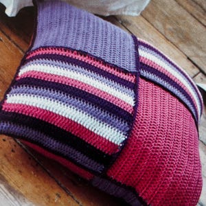 May include: A square crocheted pillow with a purple, pink, and white striped pattern on one side and a solid pink side on the other.