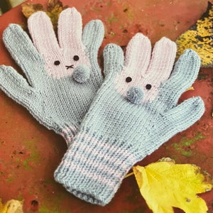 Op de afbeelding: Een paar grijze en roze gebreide konijnen wanten met zwarte ogen en een witte pompon neus.